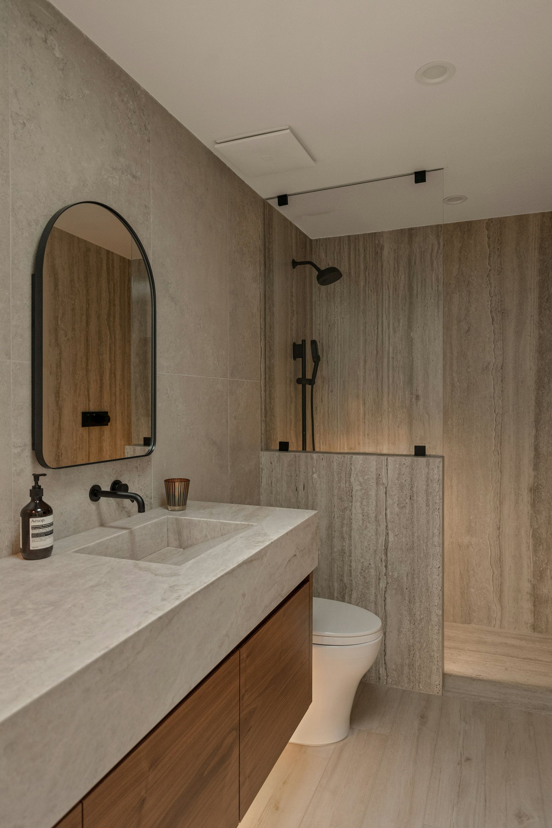 Salle de bains en pierre claire, meuble en bois, douche à l’italienne derrière un muret